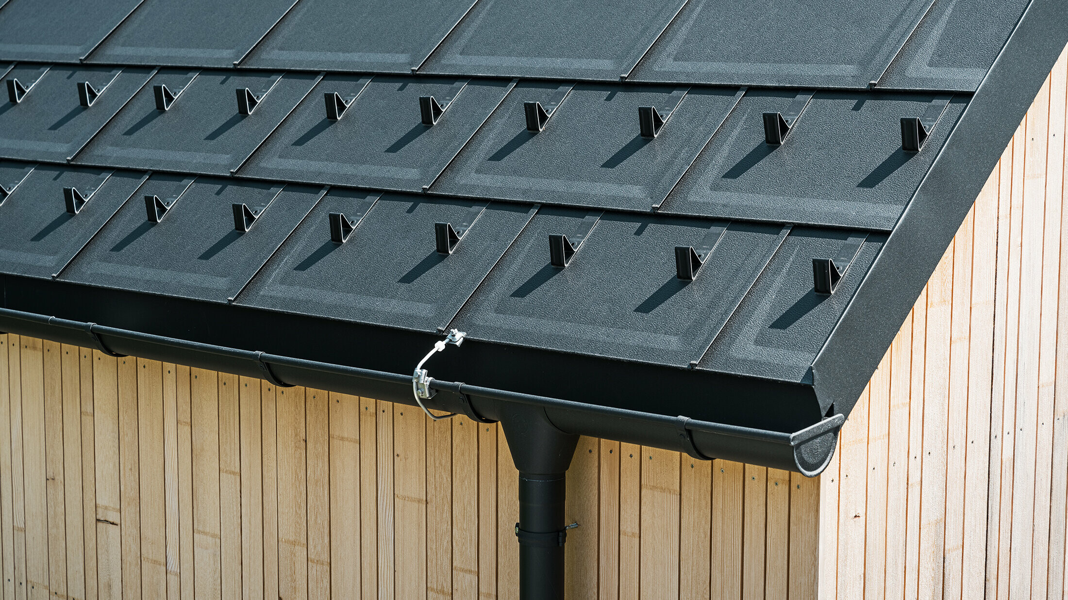 Close-up of PREFA roof tile R.16 with snow guards, gutter and downpipe in black P.10 incl. lightning protection and a vertical wooden façade
