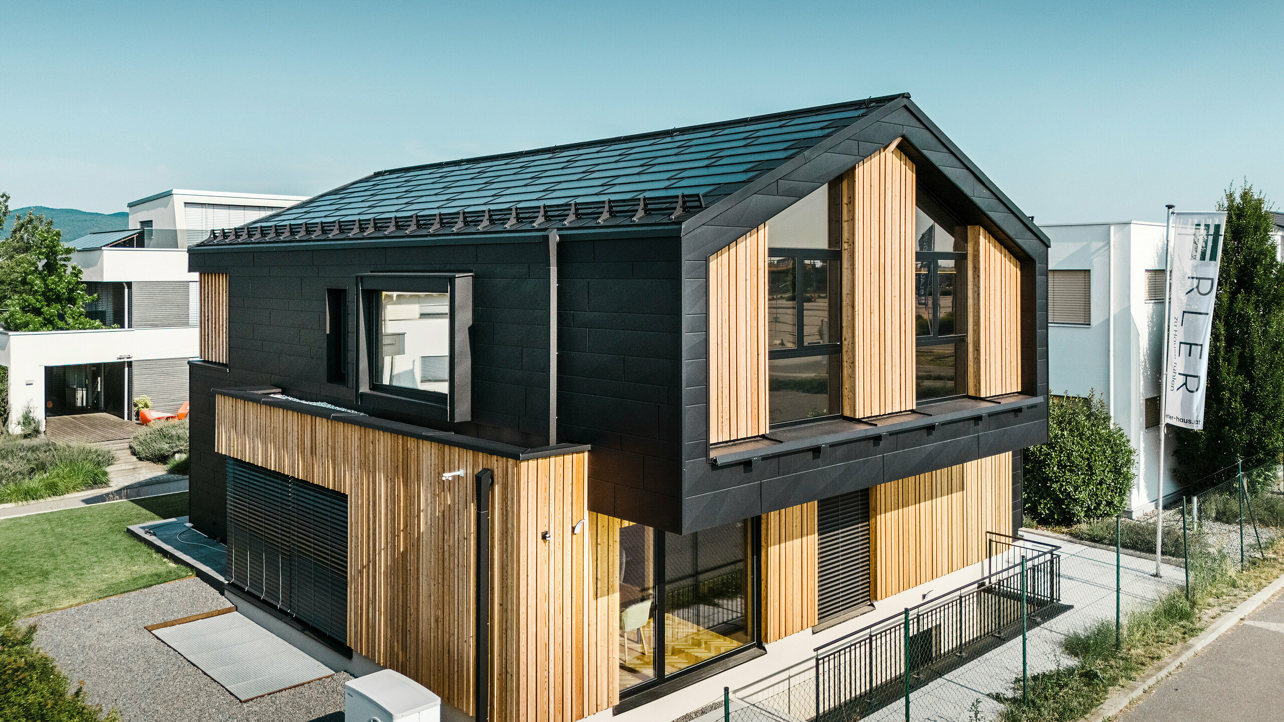 Front of the Autark show house at the Blaue Lagune prefabricated house centre in Vösendorf (Austria). The roof is covered with black PREFA solar roof panels and R.16 roof tiles with a high-quality P.10 coating. The façade consists of a combination of vertical wooden elements and PREFA siding.X in P.10 black, which gives the building a modern and sustainable character. The Erler Bau GmbH flag is flying on the right-hand side of the picture.