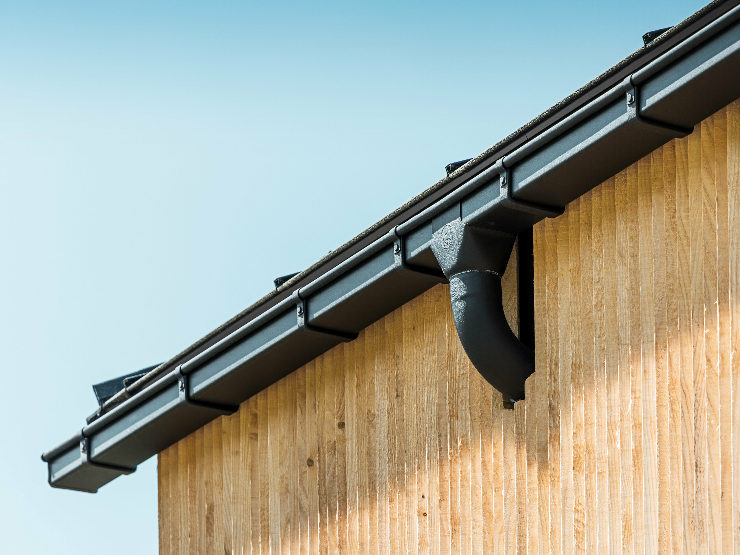 House corner with PREFA box gutter in black, with gutter box, fastened with PREFA gutter brackets, façade clad with wood.