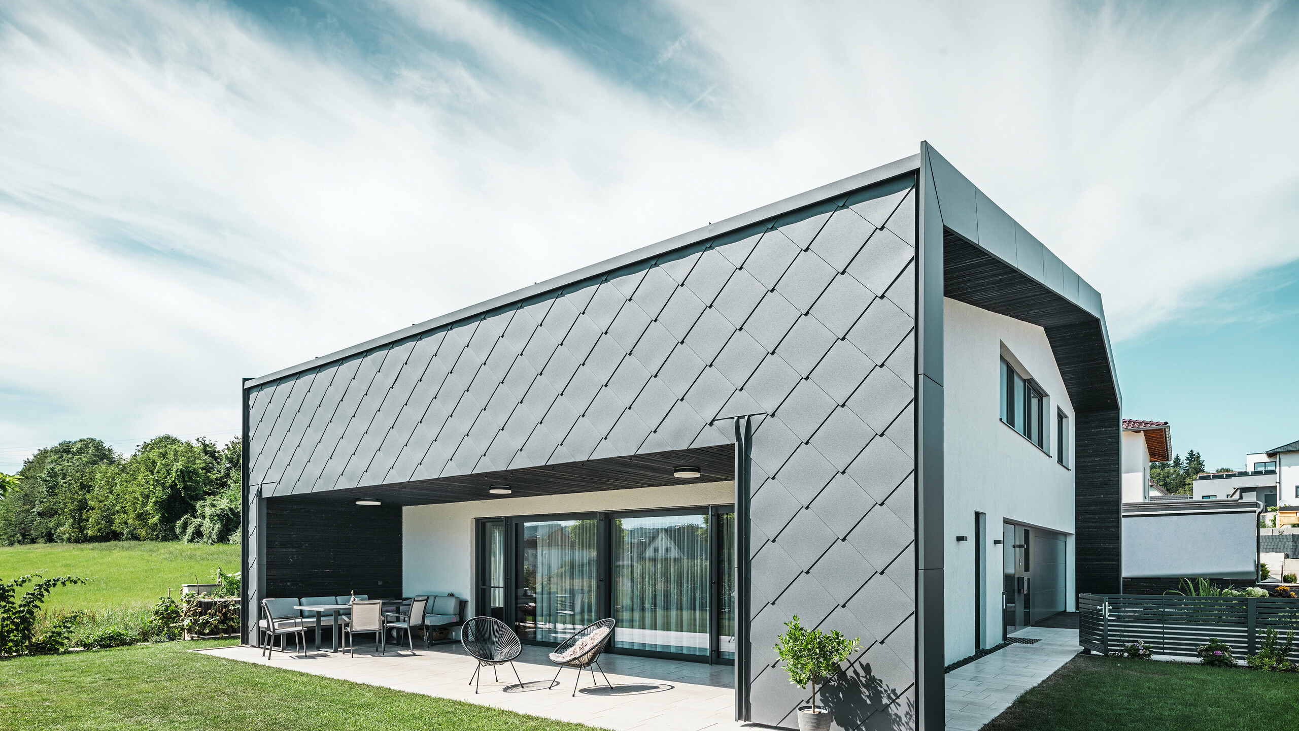 Well-kept garden area of a detached house in Aschach. The building is stylishly clad with dark grey PREFA aluminium rhomboid façade tiles 44 × 44. The covered terrace offers a cozy place to sit with a view of the lush green lawn. The elegant aluminium cladding in combination with white plaster surfaces ensures a modern and weather-resistant architecture. Large windows allow plenty of daylight into the interior, creating an open, light-filled atmosphere.
