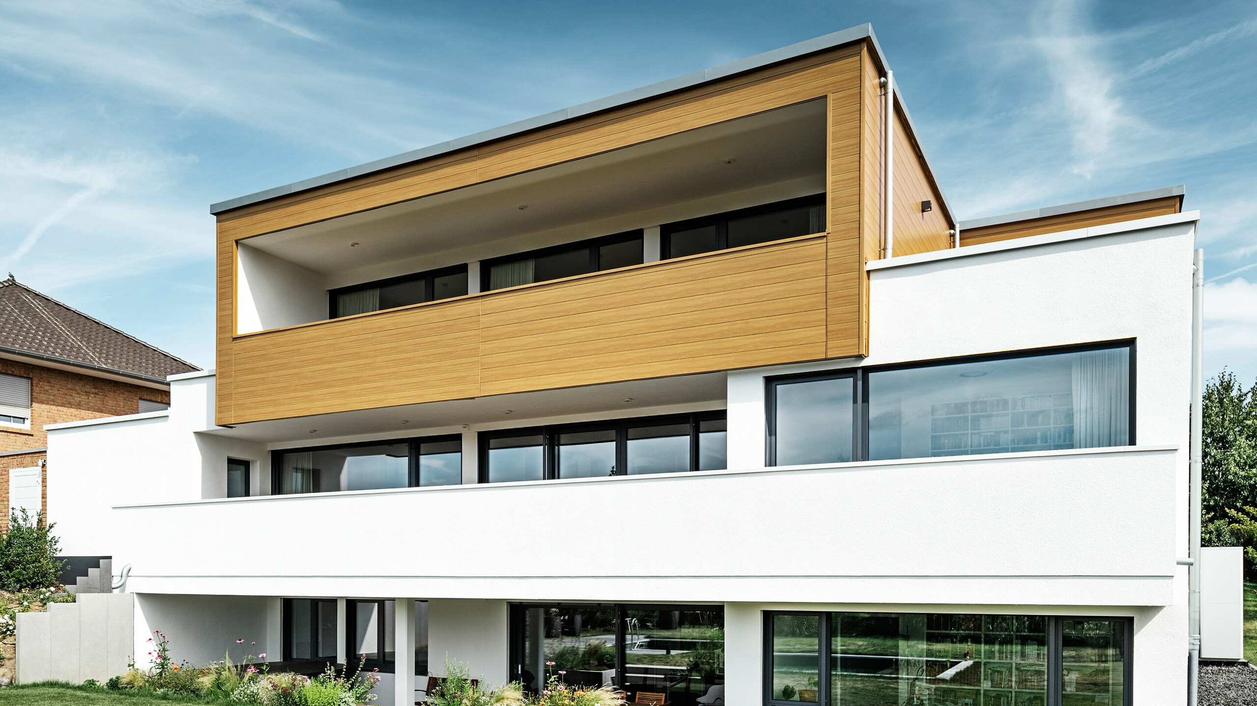 The rear of a modern detached house in Usingen, Germany. The green lawn, a pool and a young garden tree can be seen at the bottom of the picture. The eye-catching house has a white building base made of a rendered façade, an aluminium façade on the upper floor made of PREFA sidings in natural oak and large window areas with an anthracite-coloured frame. From a distance, the neighbouring house with a brick façade can be seen on the left edge of the picture.