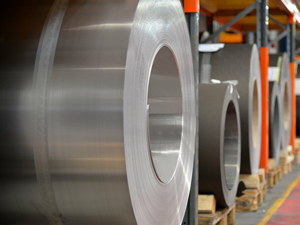 Aluminum coils stored on wooden pallets, with a plain aluminum coil in the foreground and further aluminum coils and sheets behind it.