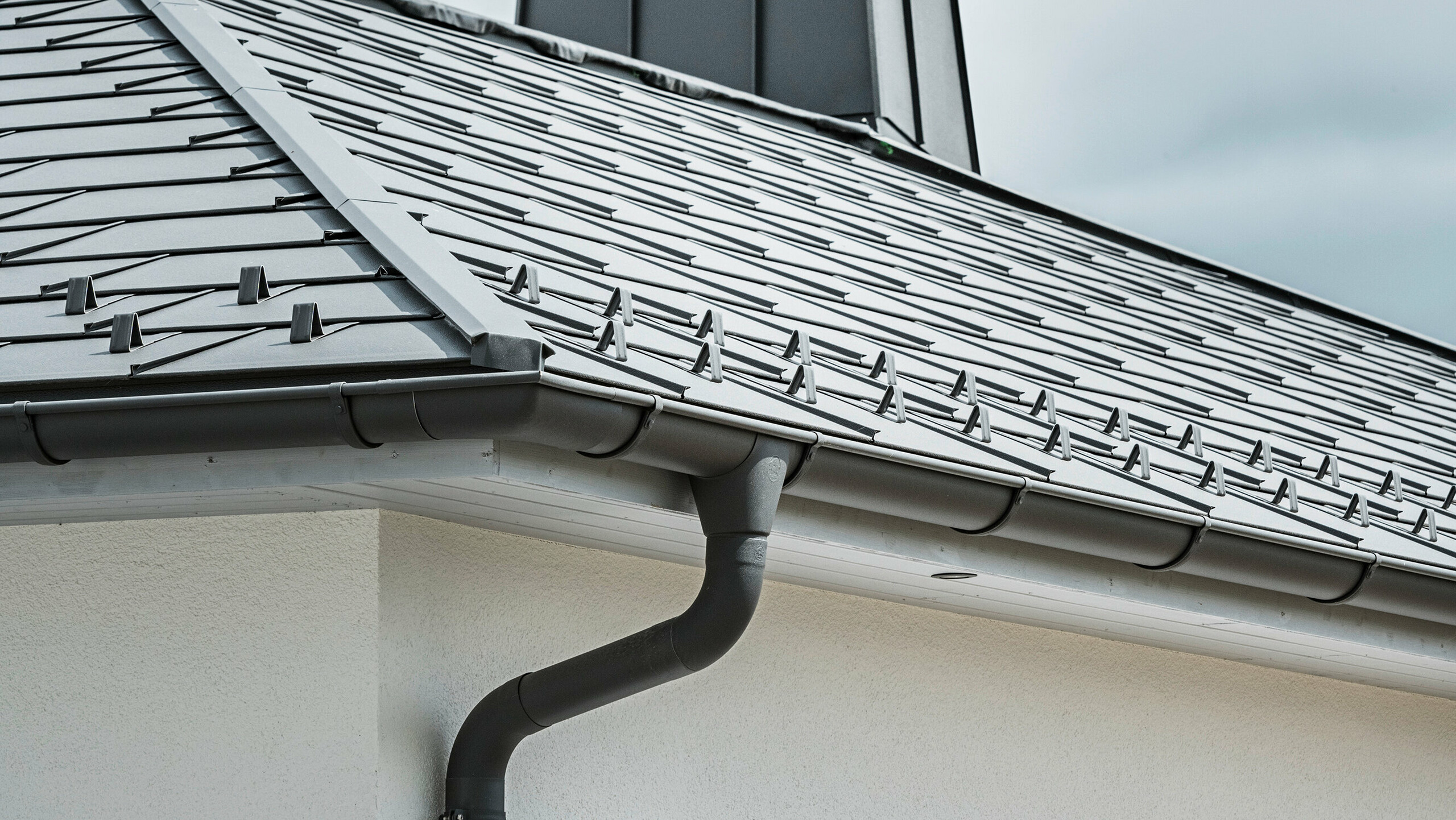 Close-up of the eaves of a detached house equipped with the efficient PREFA roof drainage system in P.10 dark grey. The image shows the detailed installation of the PREFA R.16 roof tile with the seamlessly integrated half-round gutter and downpipe underneath, which together provide a reliable and aesthetically pleasing solution for rainwater drainage. The robust aluminium products and careful installation underline the quality and durability of the PREFA complete system.