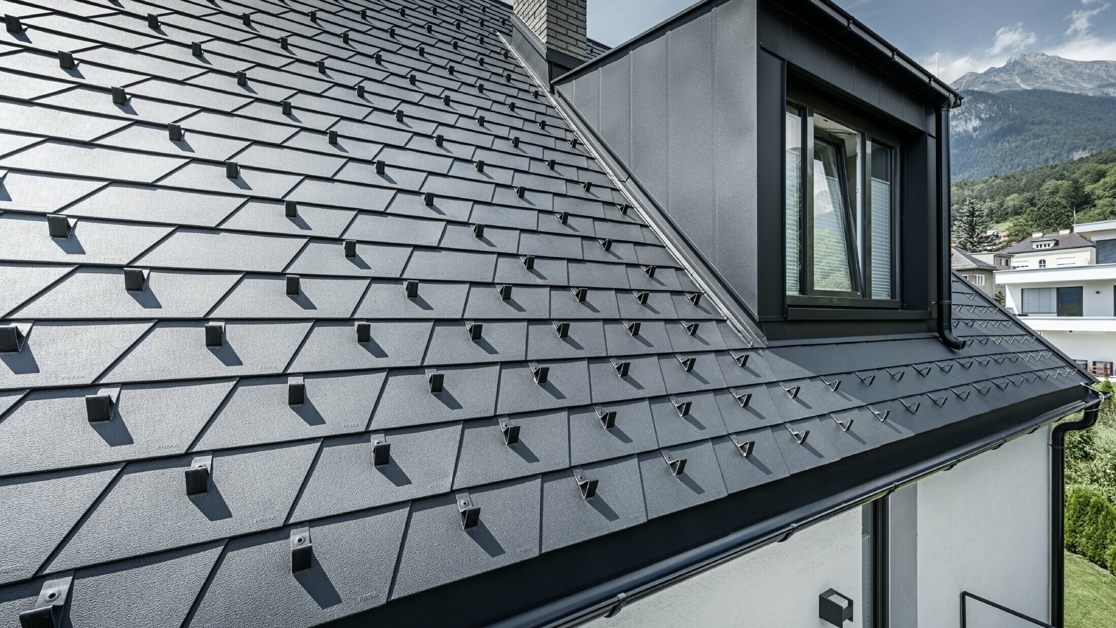 Detailed view of a roof with dormer and window, PREFA roof shingles in black with snow guards are laid on the roof surface.