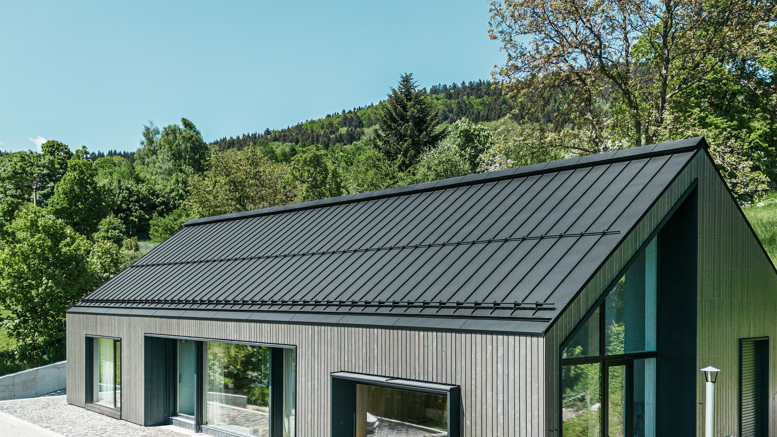 A modern wooden house in Kuncice (CZ) was equipped with large windows. The dark wooden house has a PREFALZ (standing seam) roof system in the colour P.10 black. In the rear outdoor area, there is a hot tub with wooden panelling.