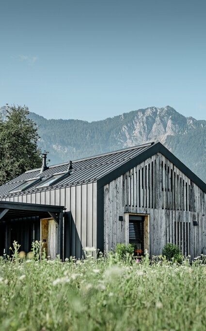 Moderner, funktionaler Wohnbau mit einer Kombination aus Holzfassade und PREFA Band- und Blechmaterial für das Dach. Die klare Architektur wird durch die elegante, metallische Dach- und Fassadenverkleidung unterstrichen, die ideal für anspruchsvolle Projekte ist. Das Bild veranschaulicht die vielseitigen Anwendungsmöglichkeiten der PREFA Produkte im Bereich Bänder und Bleche.