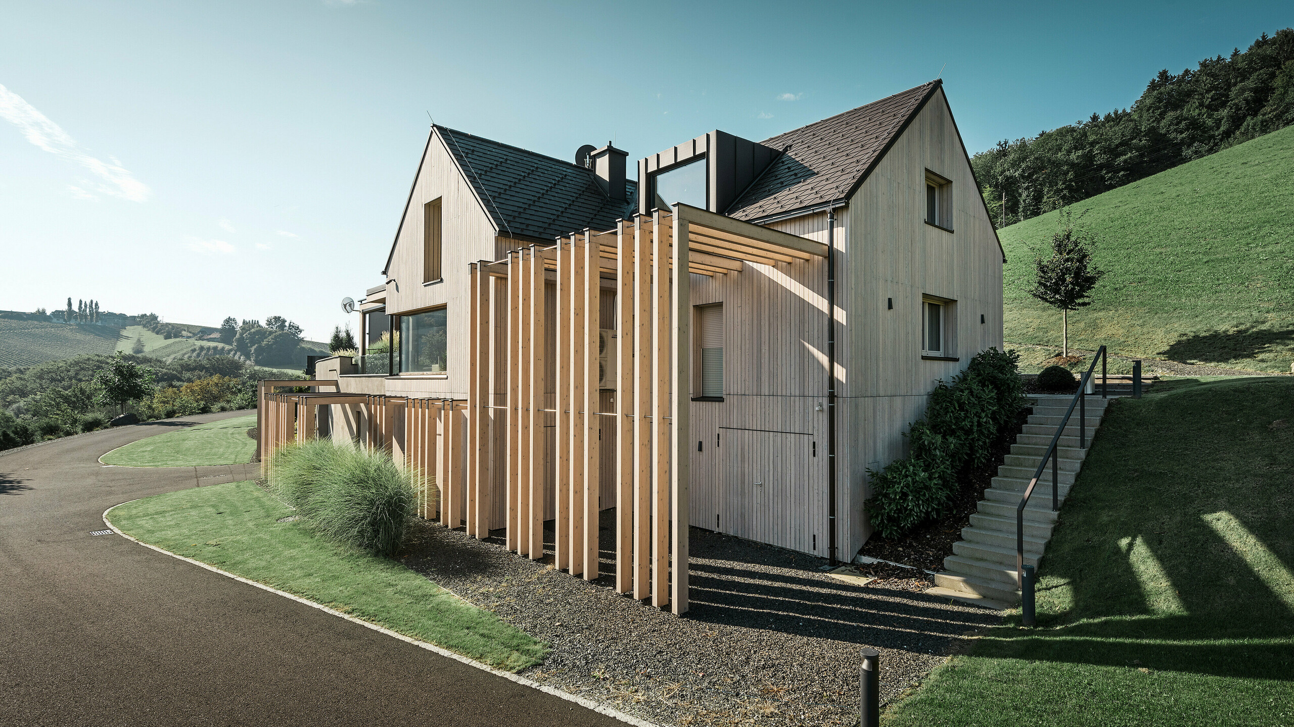 Large detached house with gabled roof and dormer window on a hillside. The roof is covered with brown PREFA aluminium shingles and the façade is clad with vertical wooden boards.