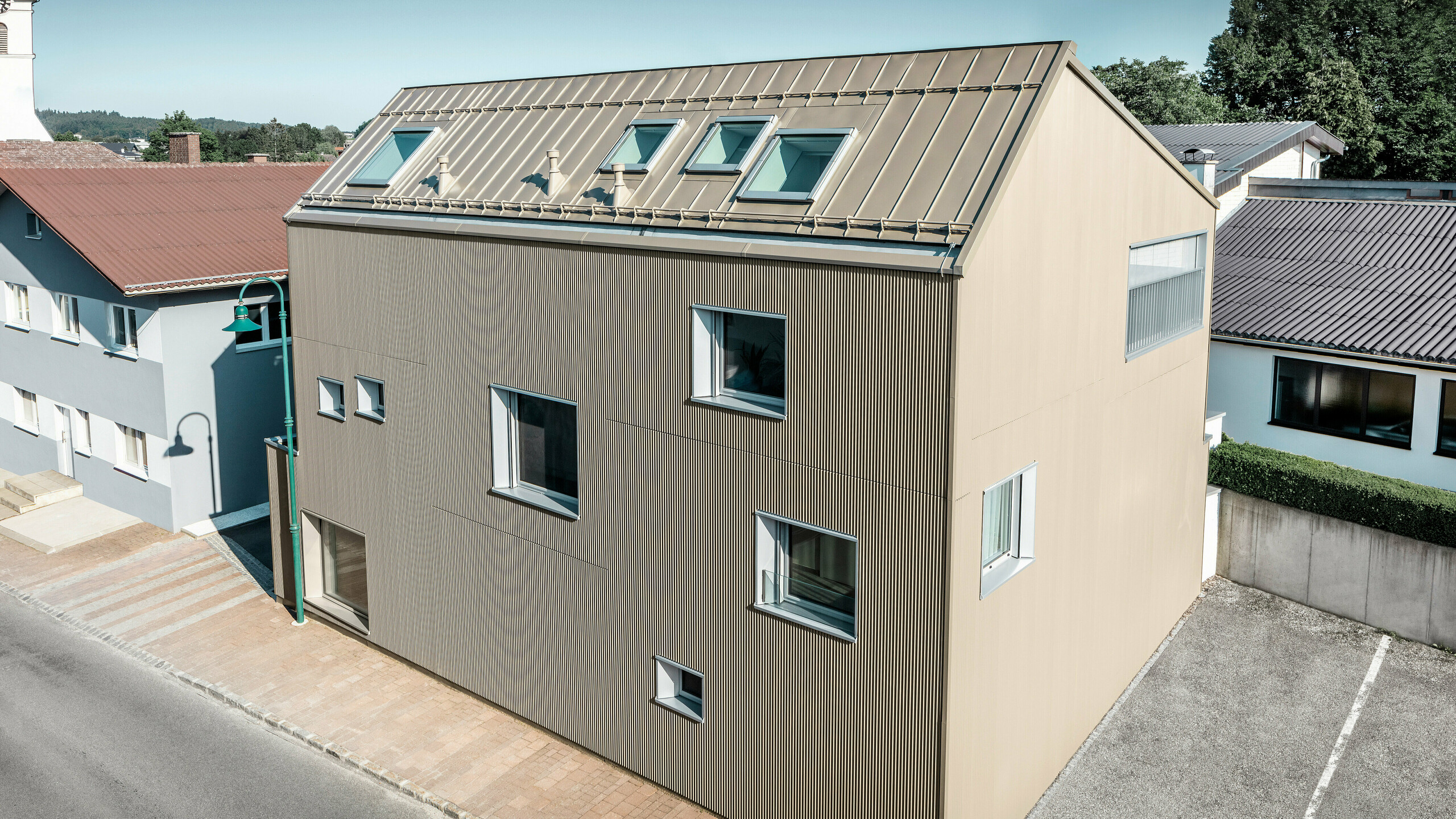 Bird's-eye view of a detached house in Frankenburg, Austria, which stands out thanks to its PREFA façade in the form of a serrated profile and a PREFALZ roof, both in a matching bronze colour. The façade has a striking texture that captures natural light and creates a lively play of shadows. Several roof windows allow natural light to flood into the internal space and emphasise the functional and aesthetic integration of modern architectural elements. With its contemporary design, the house blends harmoniously into the typical Austrian village environment while at the same time providing a contrasting accent to the surrounding traditional buildings.