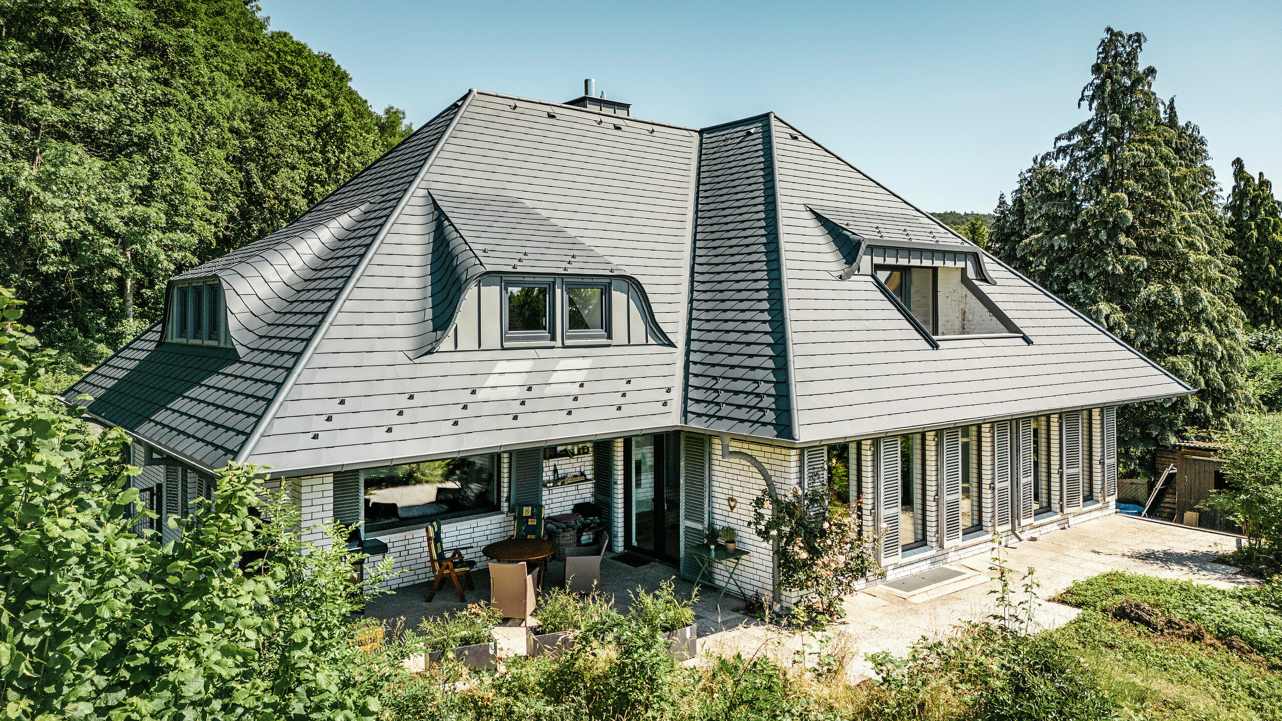 Modern detached house in Einbeck, Germany, with PREFA shingles in P.10 anthracite. The durable PREFA aluminium shingles are renowned for their quality and longevity. The roof design of the building emphasises the individual architecture with dormer windows, which are highlighted by the flexibility of the PREFA shingles. The roof drainage in the form of PREFA downpipes and half-round gutters is also in anthracite. The detached house is surrounded by a spacious and well-kept garden and a terrace. Behind the house is a forest which, together with the blue sky, creates a natural atmosphere.